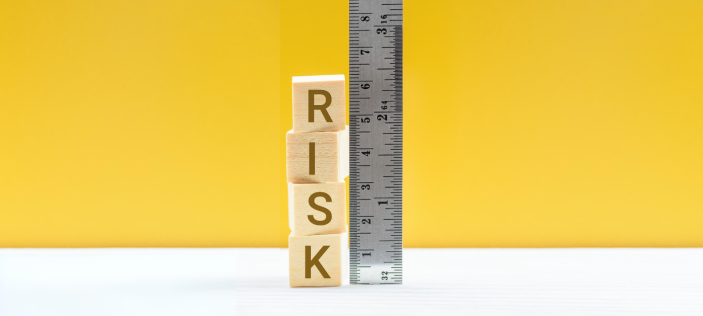 Wooden alphabet blocks spelling out 'risk' arranged vertically next to a metal ruler on a yellow and white background